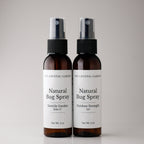 Two amber spray bottles of The Crystal Garden Natural Bug Spray standing side by side on a light gray background.