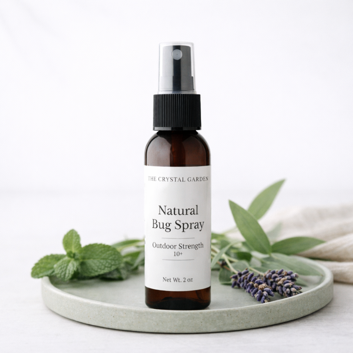 A 2 oz amber glass spray bottle of The Crystal Garden Natural Bug Spray with a white label and black spray cap, styled on a light gray ceramic tray with mint leaves and lavender sprigs against a white background.