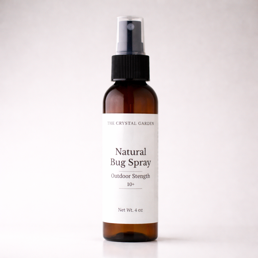 An amber glass spray bottle of The Crystal Garden Natural Bug Spray with a white label and black spray cap, set against a plain light gray background.