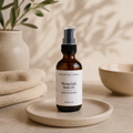 An amber glass spray bottle of 'Nourish Body Oil' by The Crystal Garden, styled on a beige ceramic tray with a neutral background and soft shadows.