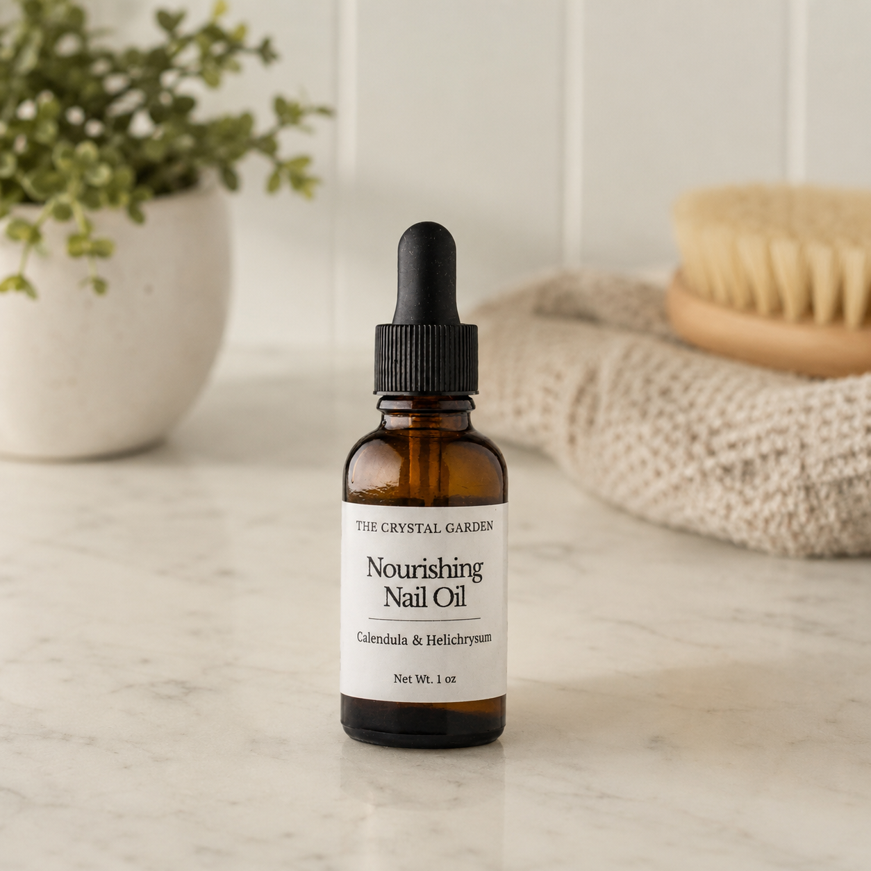 A small amber glass dropper bottle labeled 'The Crystal Garden Nourishing Nail Oil' stands on a light marble surface with a blurred bathroom-style background including a plant, towel, and brush.