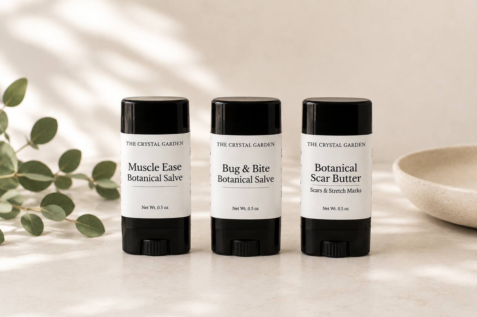 Three black and white stick containers from The Crystal Garden lined up on a light marble surface with soft leaf shadows. The products shown are Muscle Ease Botanical Salve, Bug & Bite Botanical Salve, and Botanical Scar Butter, styled with eucalyptus leaves and a ceramic bowl.