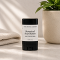 A black and white stick container of The Crystal Garden Botanical Scar Butter stands on a light marble surface with a folded beige cloth on the left and a potted green plant on the right.