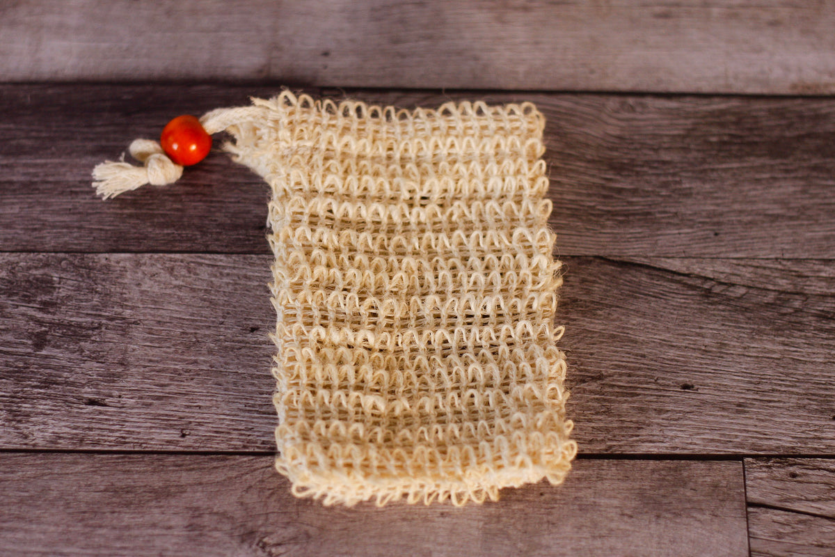 A small handmade natural fiber soap bag in a cream color, featuring a textured crochet body, a tassel tie, and a single orange-red decorative bead, displayed on a rustic dark wood background.