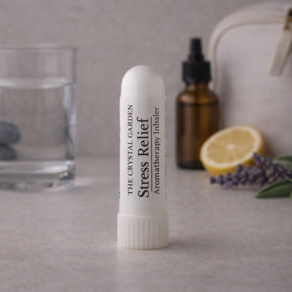 A white tube of The Crystal Garden Stress Relief Aromatherapy inhaler stands on a light gray surface, with a glass of water, amber dropper bottle, lemon slice, lavender sprigs, and a cream-colored bag blurred in the background.