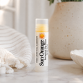 A tube of The Crystal Garden Sun Orange Botanical lip balm stands on a light marble surface, styled with a cream knit blanket, a speckled ceramic vase, and a small dish in the background.