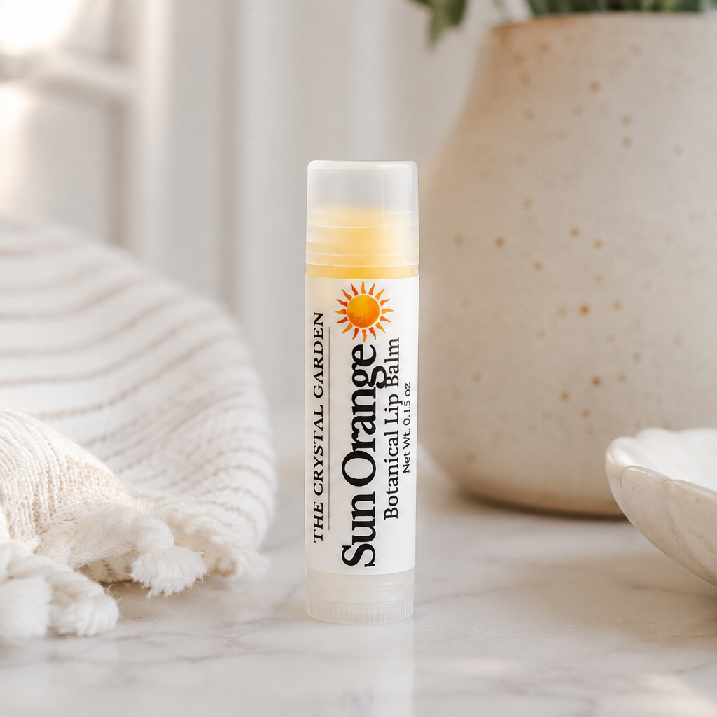 A tube of The Crystal Garden Sun Orange Botanical lip balm stands on a light marble surface, styled with a cream knit blanket, a speckled ceramic vase, and a small dish in the background.