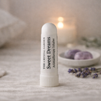 A white cylindrical aromatherapy inhaler tube labeled 'The Crystal Garden Sweet Dreams' stands on a soft neutral surface. The background is softly blurred with a warm glowing candle, lavender sprigs, and a small dish of purple stones.