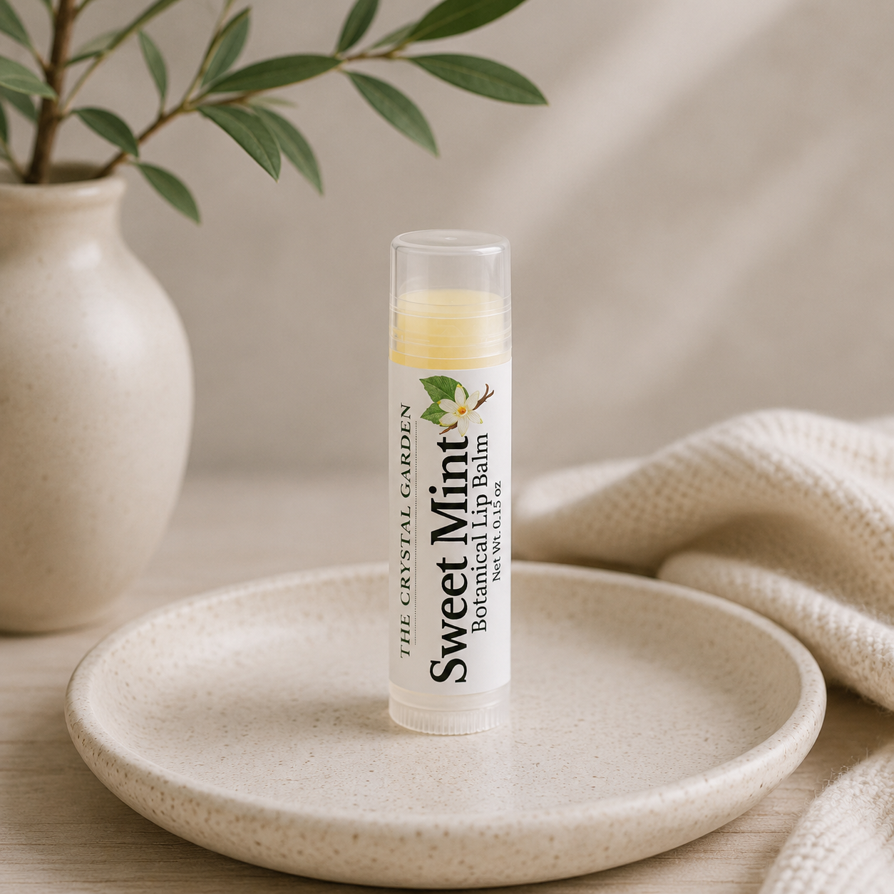 A tube of The Crystal Garden Sweet Mint balm lip balm stands on a round ceramic plate, styled with a beige knit blanket and a small potted plant in the background.
