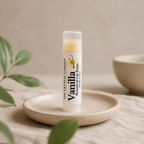 A tube of Vanilla Botanical Balm by The Crystal Garden stands upright on a small beige ceramic dish. The product packaging is white with black text and a small yellow flower illustration, and the balm inside is visible at the top.