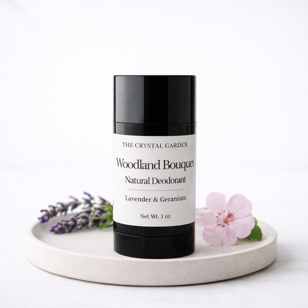 A black and white deodorant stick from The Crystal Garden, labeled Woodland Bouquet, styled on a round ceramic tray with lavender sprigs and a pink flower against a white background.