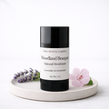 A black and white deodorant stick from The Crystal Garden, labeled Woodland Bouquet, styled on a round ceramic tray with lavender sprigs and a pink flower against a white background.