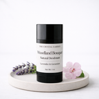 A black and white deodorant stick from The Crystal Garden, labeled Woodland Bouquet, styled on a round ceramic tray with lavender sprigs and a pink flower against a white background.