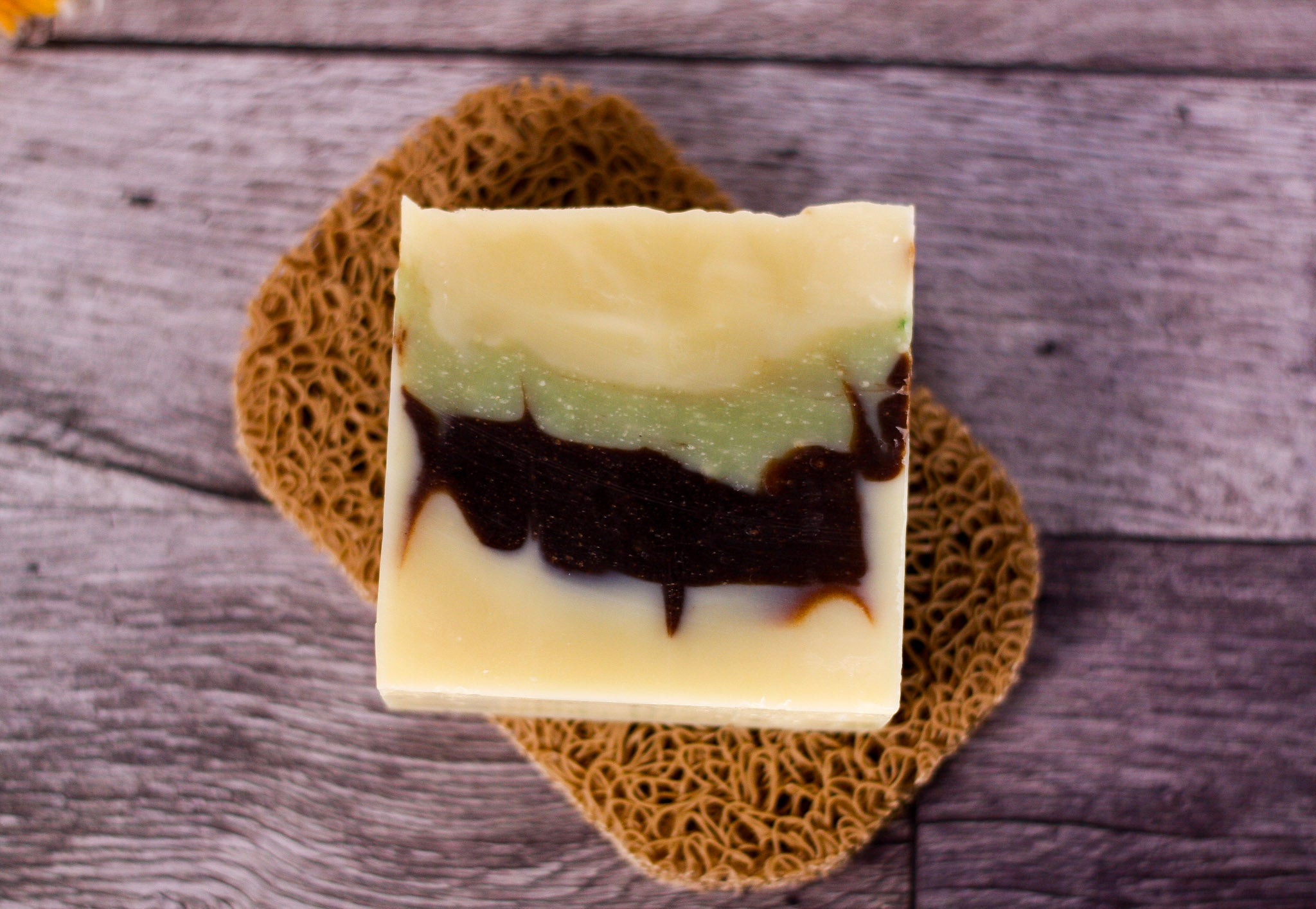 A rectangular bar of handmade soap with layered cream, light green, and dark brown swirls, displayed on a brown woven mat over a gray wooden surface.