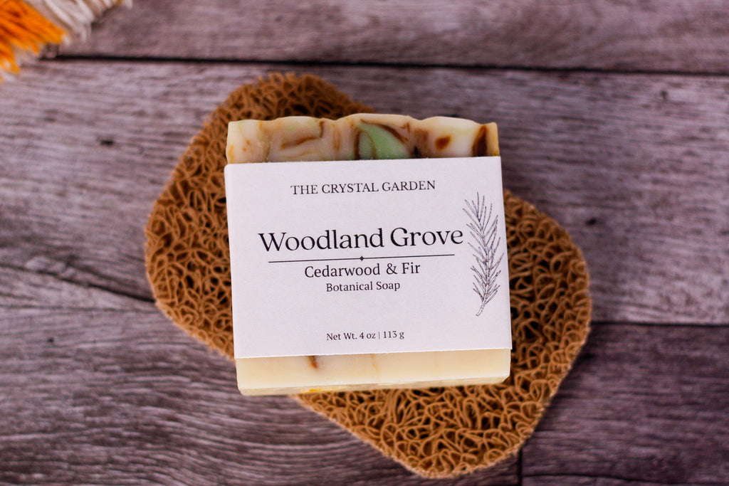 A bar of handmade soap from The Crystal Garden, named Woodland Grove, resting on a brown woven placemat over a gray wooden surface. The soap has a white label and features visible natural ingredients in shades of green, beige, and brown.