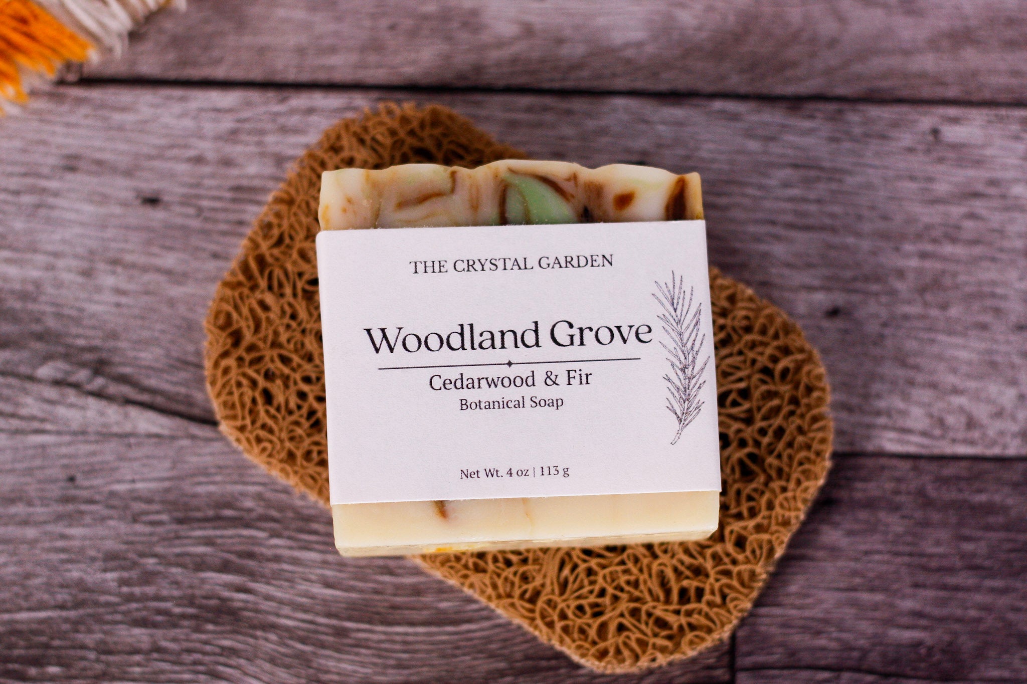 A bar of handmade soap from The Crystal Garden, named Woodland Grove, resting on a brown woven placemat over a gray wooden surface. The soap has a white label and features visible natural ingredients in shades of green, beige, and brown.