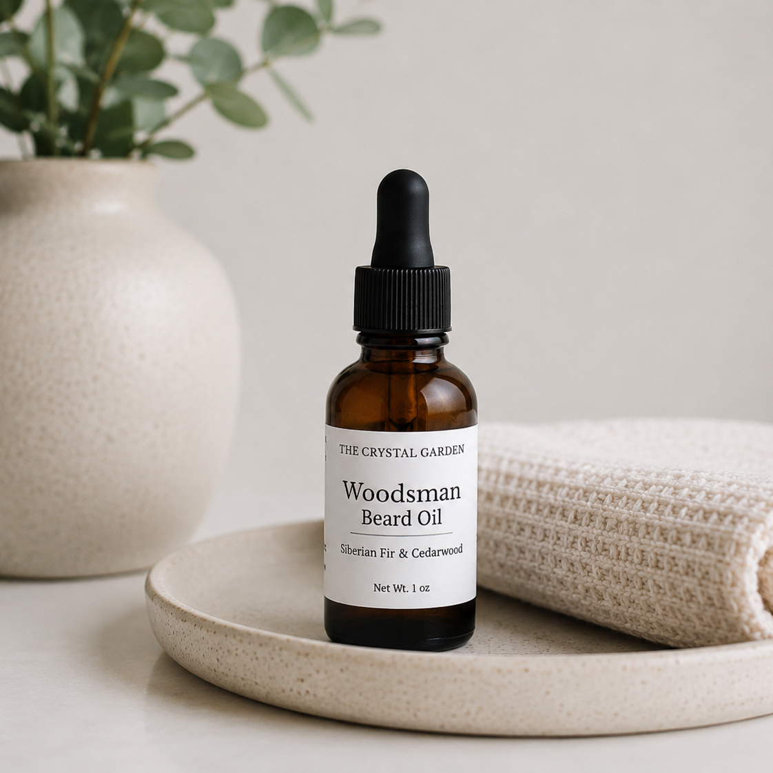 A 1 oz amber glass dropper bottle of The Crystal Garden Woodsman Beard Oil, styled on a ceramic tray next to a folded cream knit blanket and a ceramic vase with green leaves.