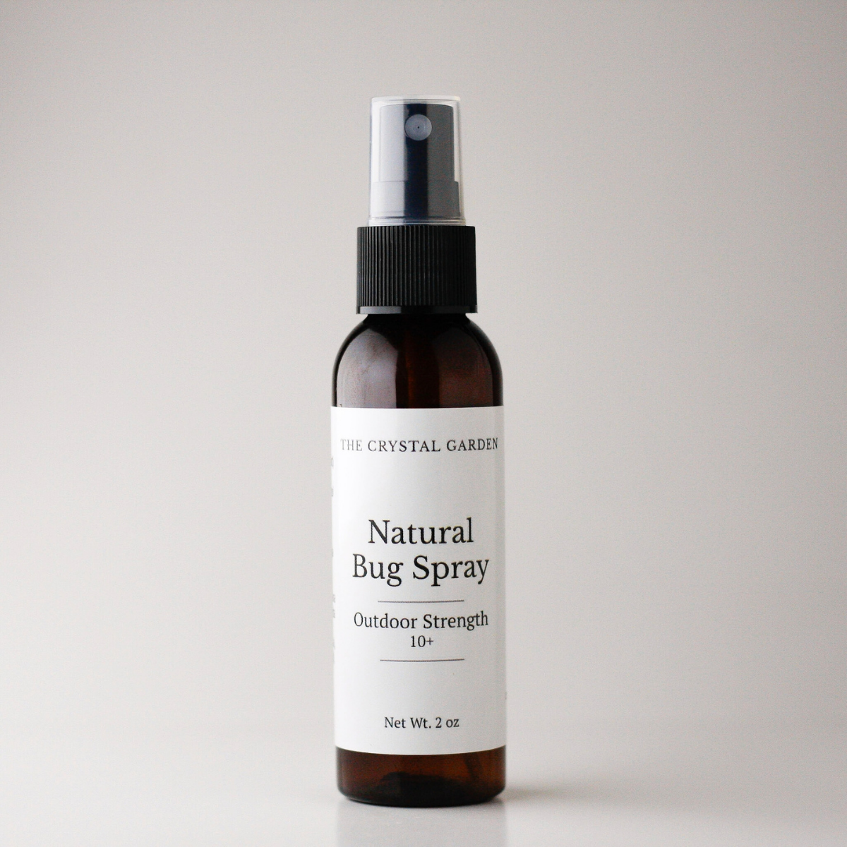 An amber glass spray bottle of 'Natural Bug Spray' by The Crystal Garden, featuring a white label and a black spray nozzle, set against a plain light gray background.