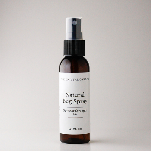 An amber glass spray bottle of 'Natural Bug Spray' by The Crystal Garden, featuring a white label and a black spray nozzle, set against a plain light gray background.