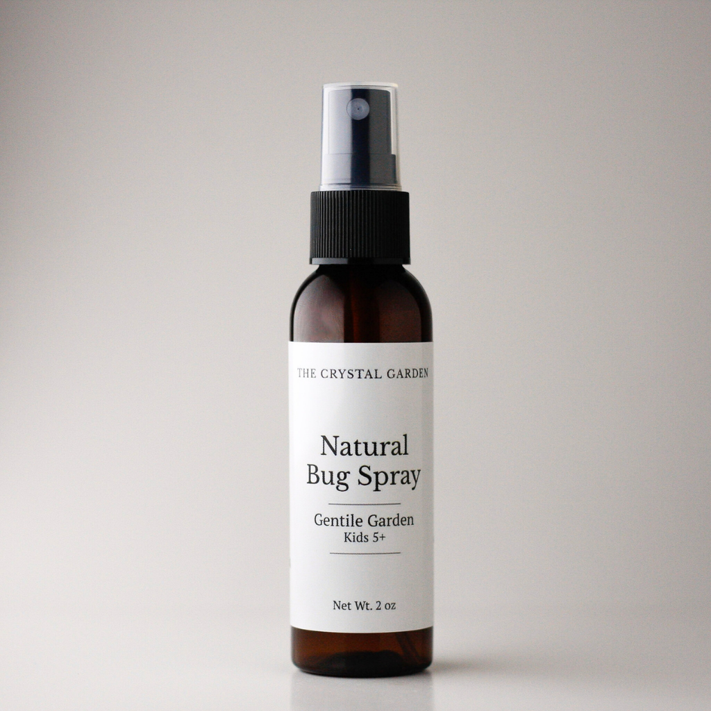 An amber glass spray bottle of The Crystal Garden Natural Bug Spray with a white label and black spray nozzle, set against a neutral light gray background.