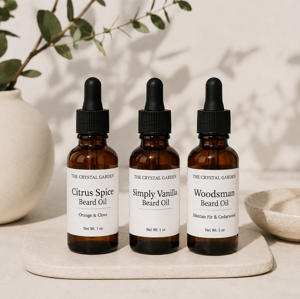 Three amber glass dropper bottles of beard oil from The Crystal Garden are lined up on a light stone tray. The bottles are labeled Citrus Spice Beard Oil, Simply Vanilla Beard Oil, and Woodsman Beard Oil, set against a warm beige background with soft shadows and a ceramic vase with green leaves.