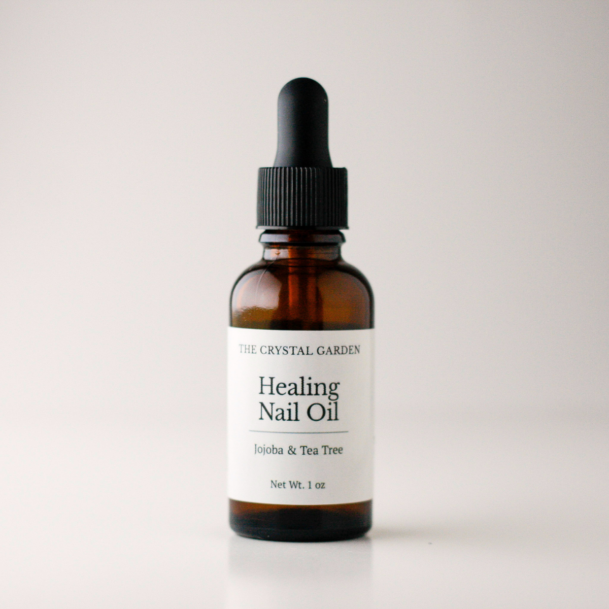 An amber glass dropper bottle of The Crystal Garden Healing Nail Oil on a light gray background.