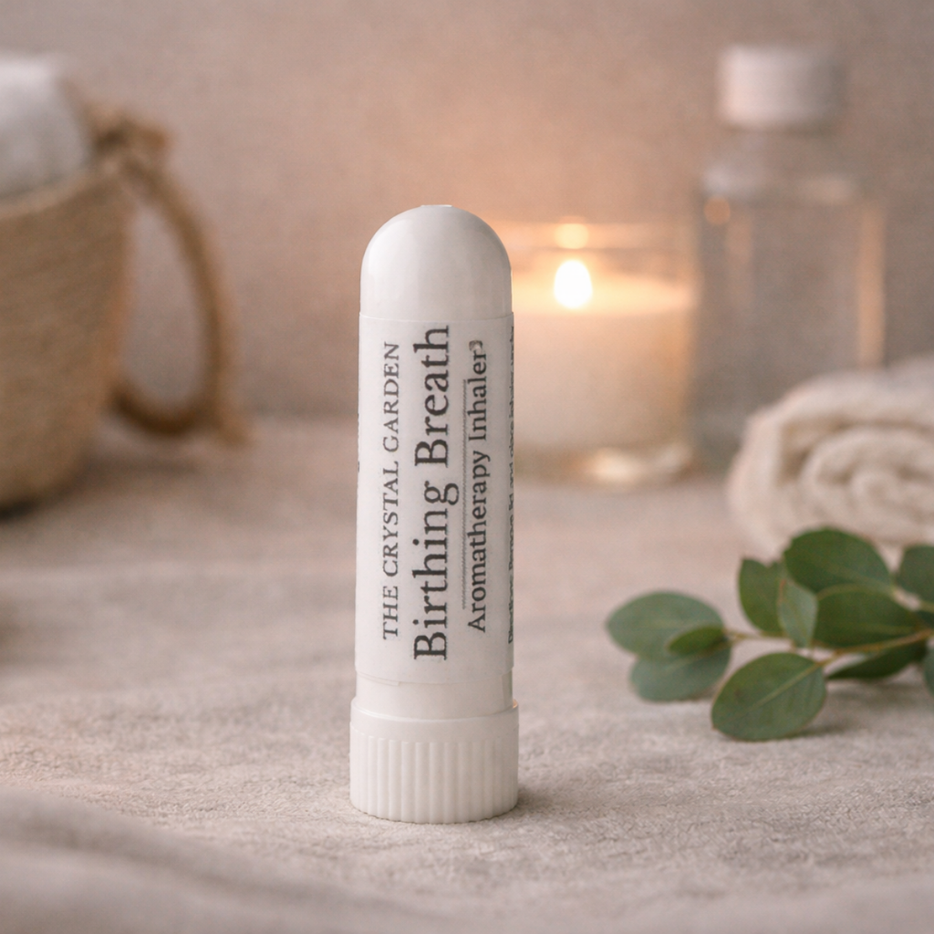 A white cylindrical balm-style inhaler labeled 'The Crystal Garden Birthing Breath' stands on a soft beige fabric surface. The background is softly blurred with a lit candle, a glass bottle, a woven basket, and green leaves, creating a spa-like setting.