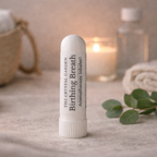 A white cylindrical balm-style inhaler labeled 'The Crystal Garden Birthing Breath' stands on a soft beige fabric surface. The background is softly blurred with a lit candle, a glass bottle, a woven basket, and green leaves, creating a spa-like setting.