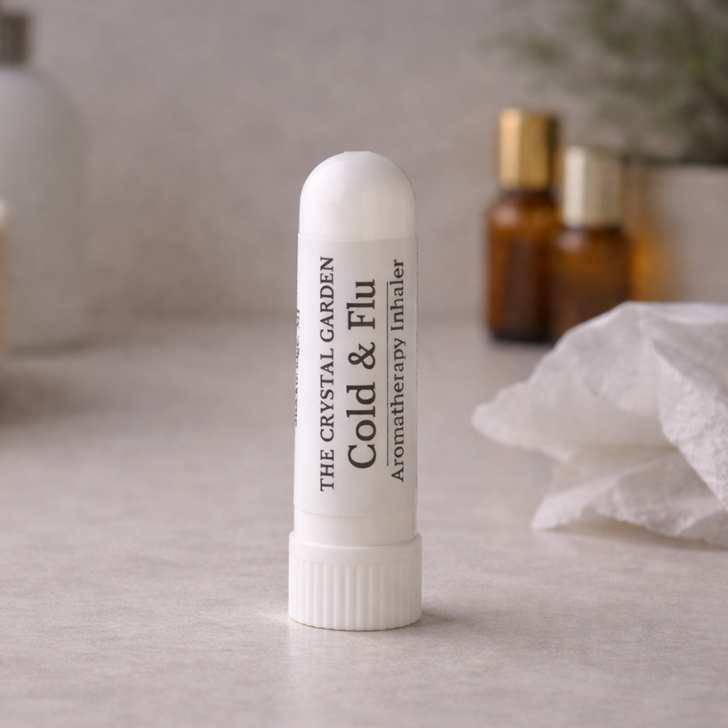 A white cylindrical cold and flu aromatherapy inhaler tube by The Crystal Garden, standing upright on a light gray surface with blurred amber bottles and a tissue in the background.