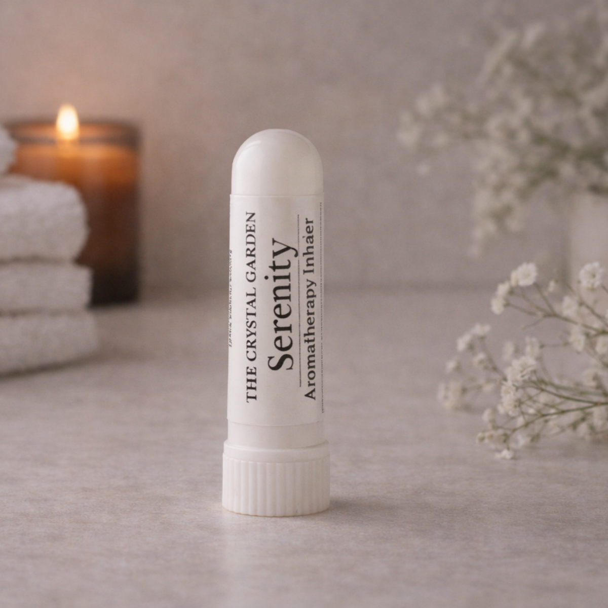 A white cylindrical aromatherapy inhaler tube labeled 'Serenity' by The Crystal Garden, standing on a light stone surface with a softly blurred candle, folded towels, and small white flowers in the background.