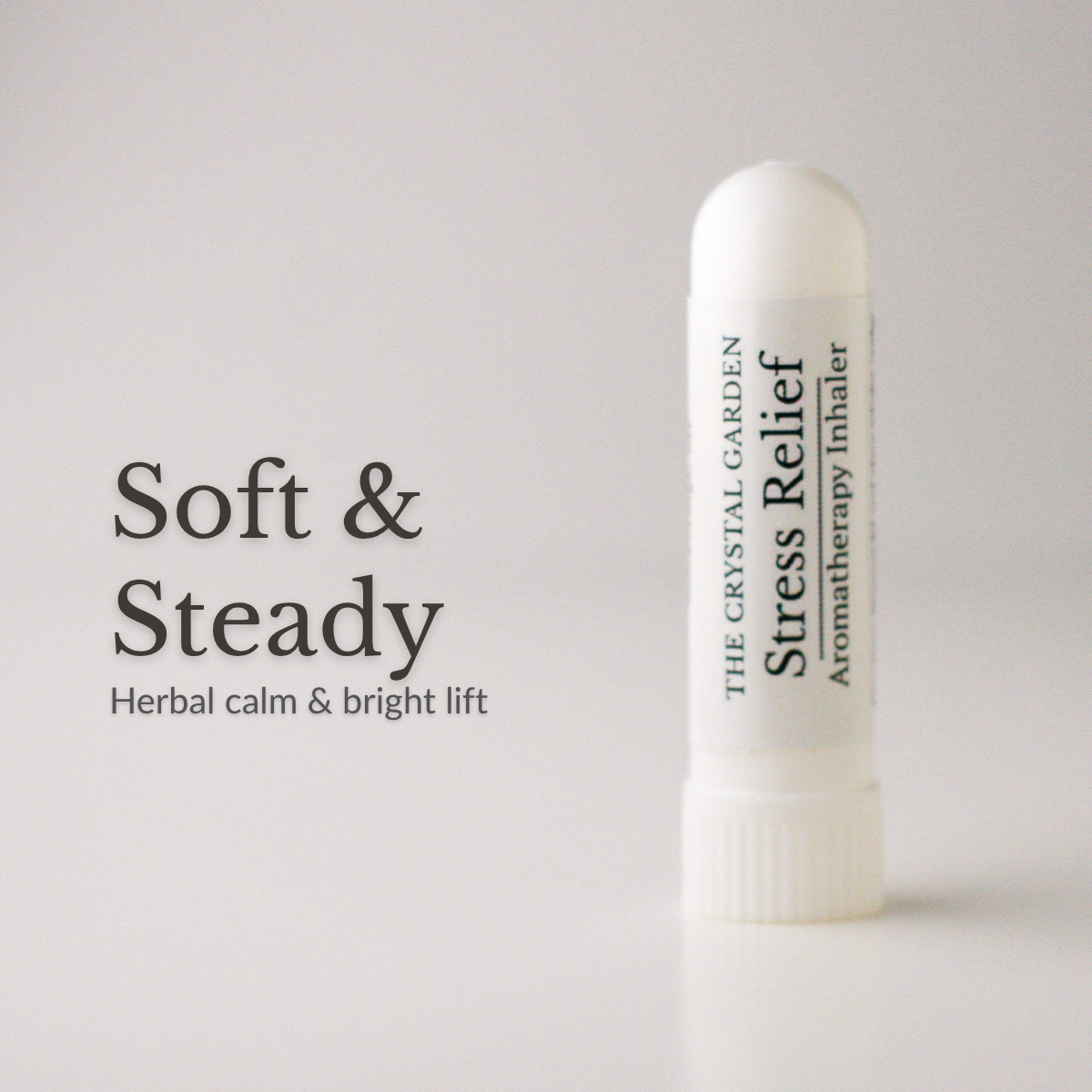 A white cylindrical stress relief aromatherapy inhaler by The Crystal Garden stands on a light gray surface against a light gray background. To the left, large black text reads 'Soft & Steady' with smaller text 'Herbal calm & bright lift'.