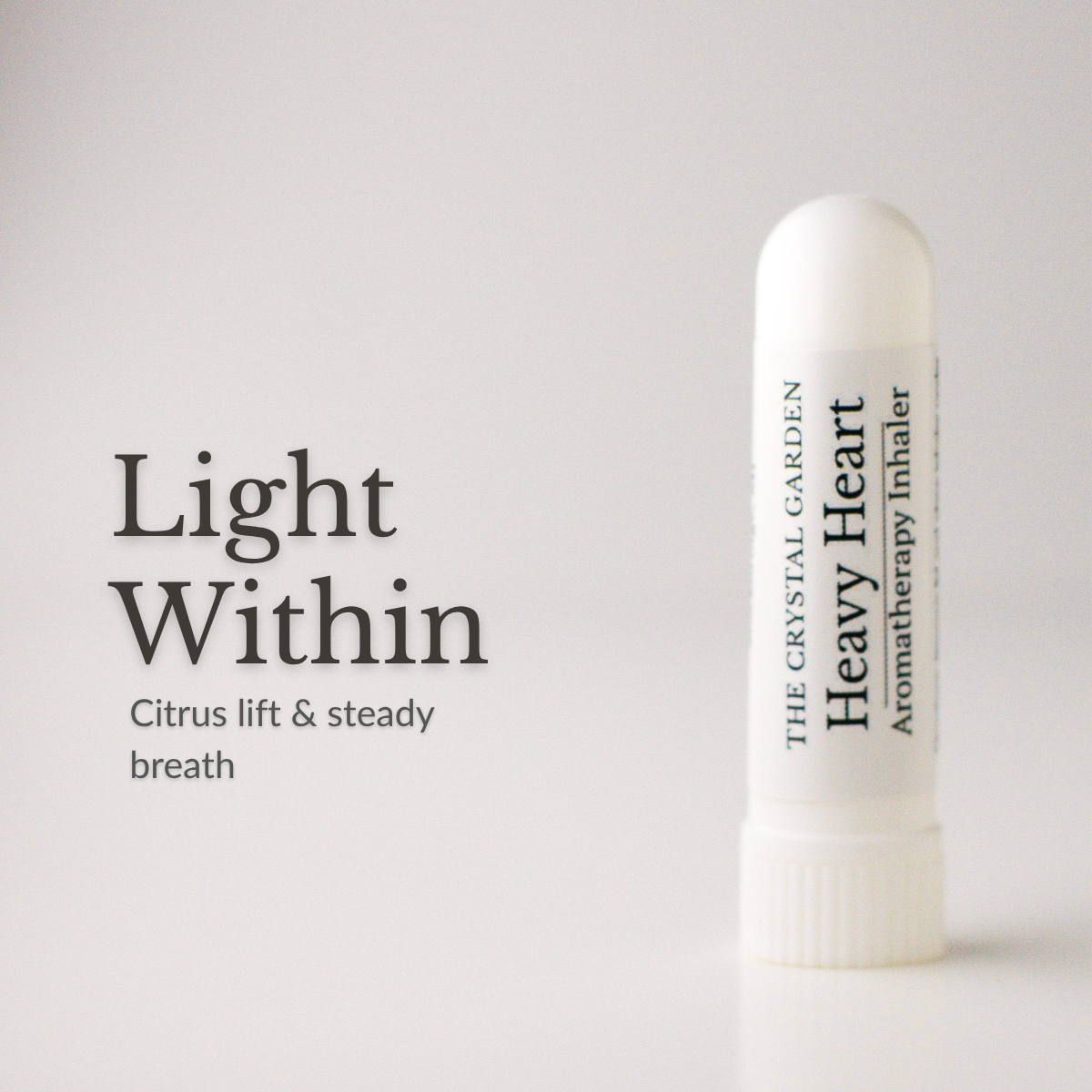 A white cylindrical aromatherapy inhaler tube labeled 'THE CRYSTAL GARDEN Heavy Heart Aromatherapy Inhaler' stands on a light gray background beside the text 'Light Within' and 'Citrus lift & steady breath'.