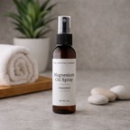 A bottle of The Crystal Gardens Magnesium Oil Spray, Unscented, standing on a gray stone surface with a rolled white towel, a potted plant, and smooth stones in the background.