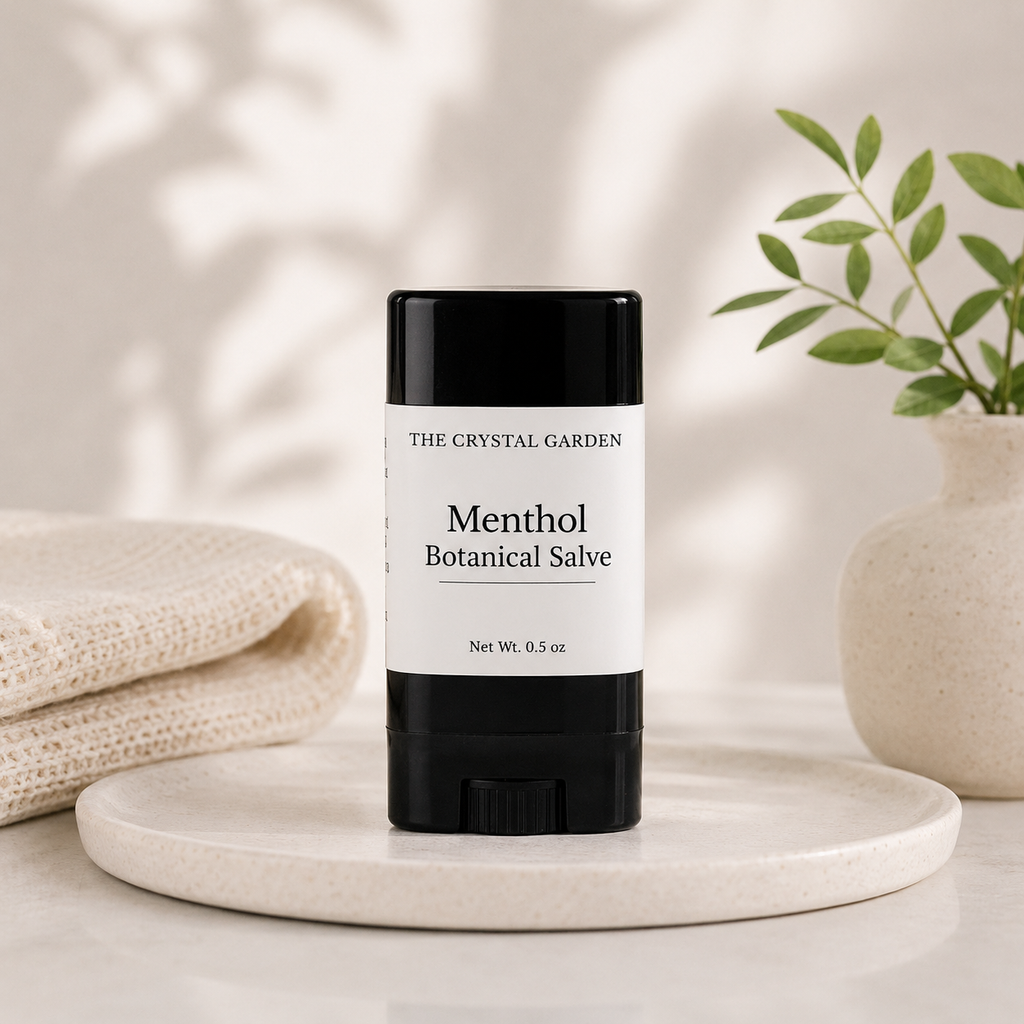 A black and white stick of The Crystal Garden Menthol Botanical Salve sits on a round marble tray, styled with a folded cream knit blanket on the left and a small potted plant on the right.
