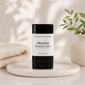 A black and white stick of The Crystal Garden Menthol Botanical Salve sits on a round marble tray, styled with a folded cream knit blanket on the left and a small potted plant on the right.