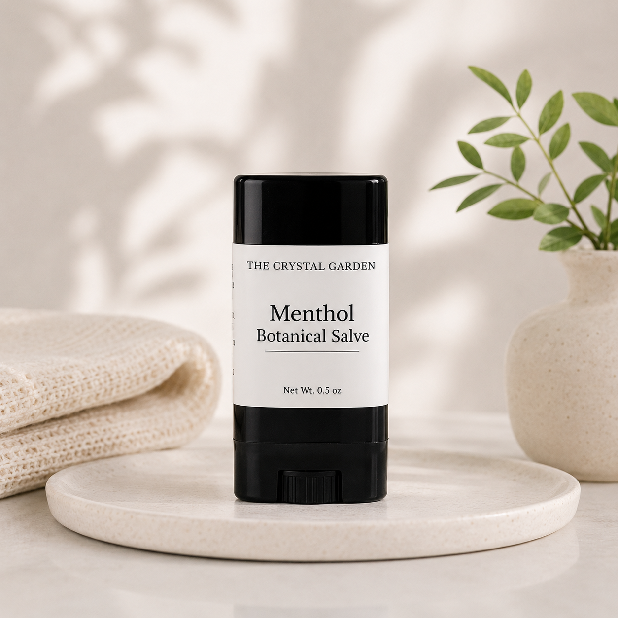 A black and white stick of The Crystal Garden Menthol Botanical Salve sits on a round marble tray, styled with a folded cream knit blanket on the left and a small potted plant on the right.