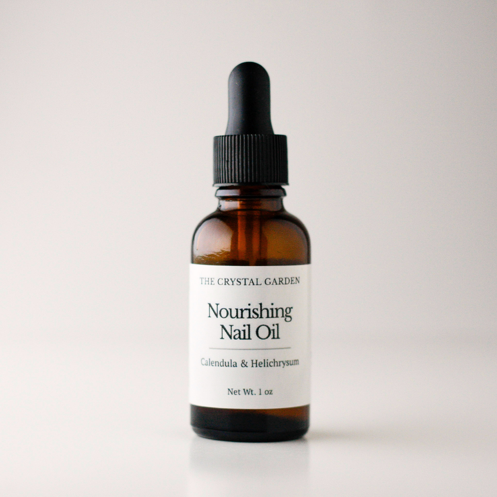 An amber glass dropper bottle of The Crystal Garden Nourishing Nail Oil with a white label and black dropper cap, set against a clean light gray background.