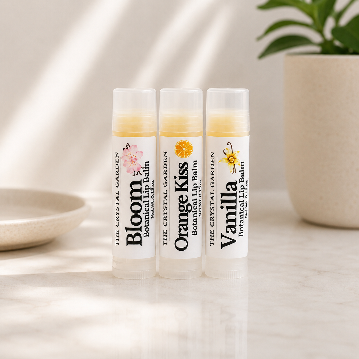 Three lip balm tubes standing upright on a light marble surface, with a beige ceramic planter and plant in the background. The tubes are labeled Bloom, Orange Kiss, and Vanilla Botanical Lip Balm by The Crystal Garden.