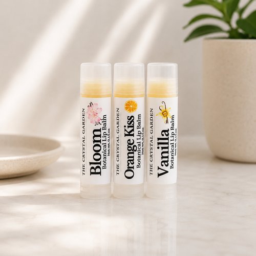 Three lip balm tubes standing upright on a light marble surface, with a beige ceramic planter and plant in the background. The tubes are labeled Bloom, Orange Kiss, and Vanilla Botanical Lip Balm by The Crystal Garden.