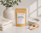 A stand-up kraft pouch labeled 'The Crystal Garden Botanical Mineral Soak Restore Single Use' stands on a light marble surface with a small bowl of white powder, a wooden scoop, folded cream towels, and a ceramic vase with greenery in the background.
