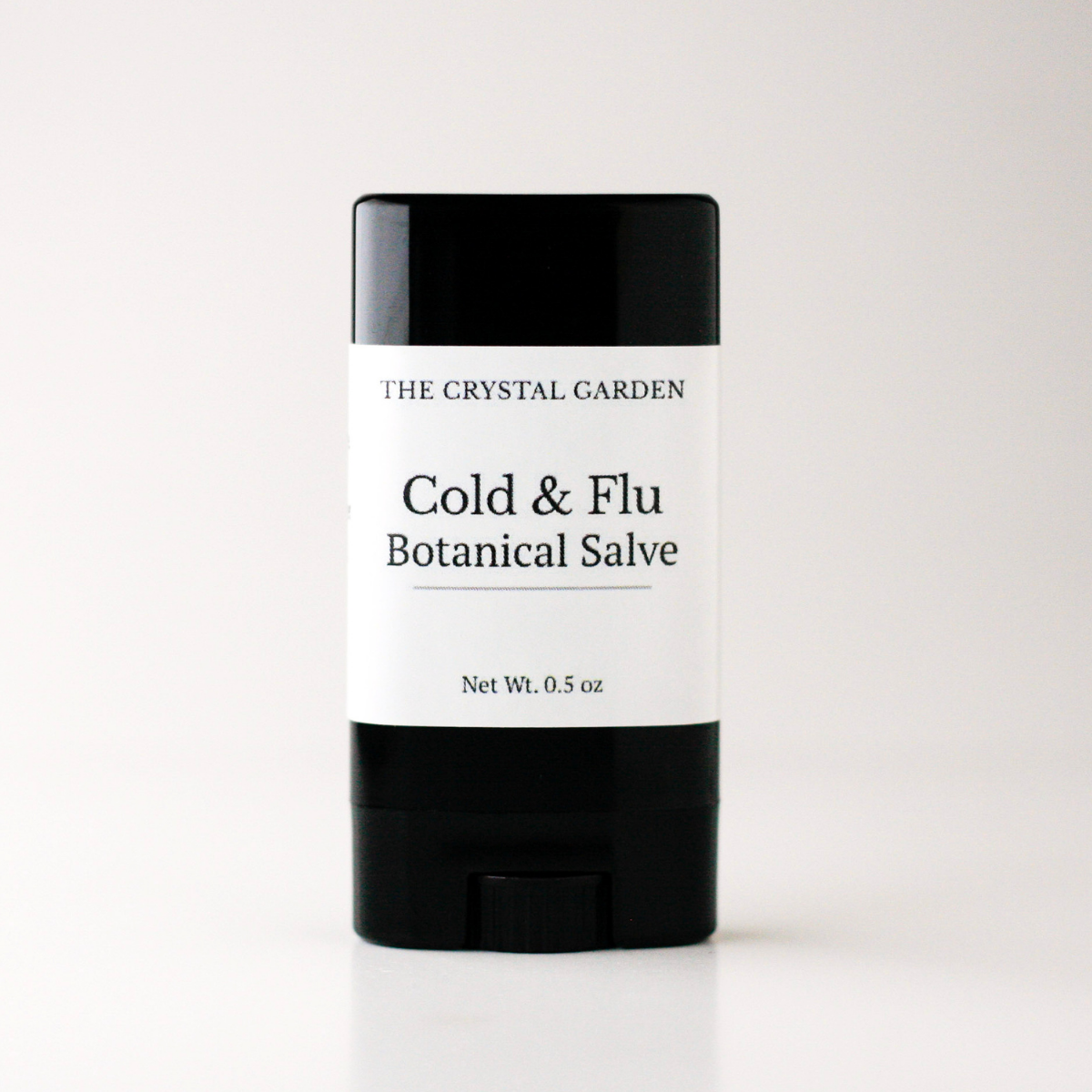 A single stick of salve from The Crystal Garden, Cold & Flu Botanical Salve, in a black and white container, centered on a light gray background.