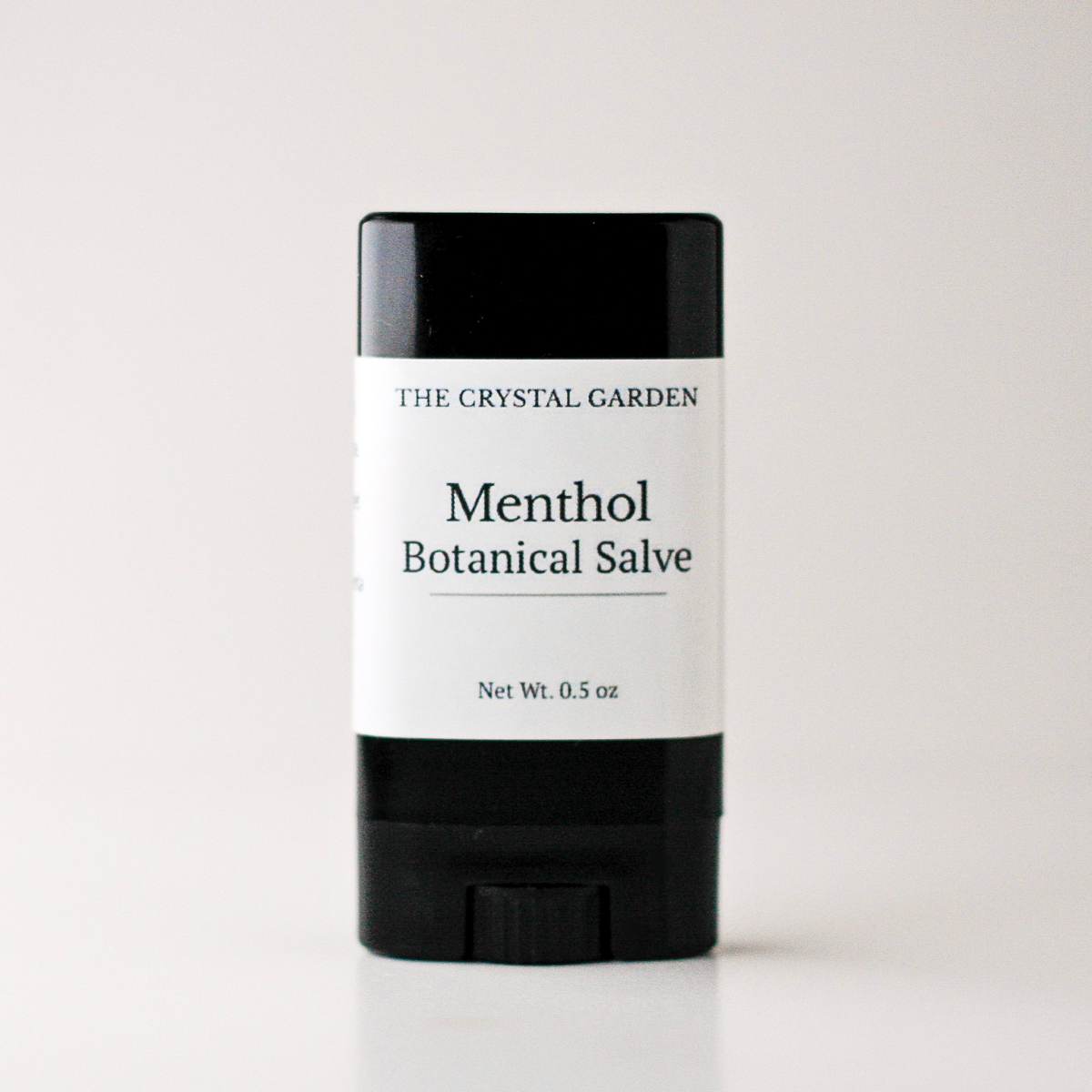 A single stick of The Crystal Garden Menthol Botanical Salve, featuring a black container with a white label, centered on a light gray background.