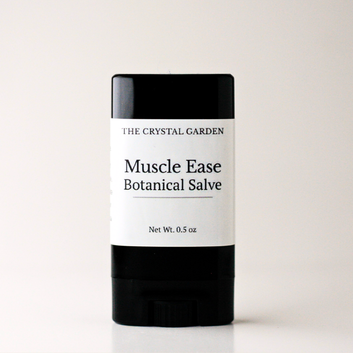 A single stick of 'Muscle Ease Botanical Salve' by 'The Crystal Garden' in a black container with a white label, centered on a light gray background.