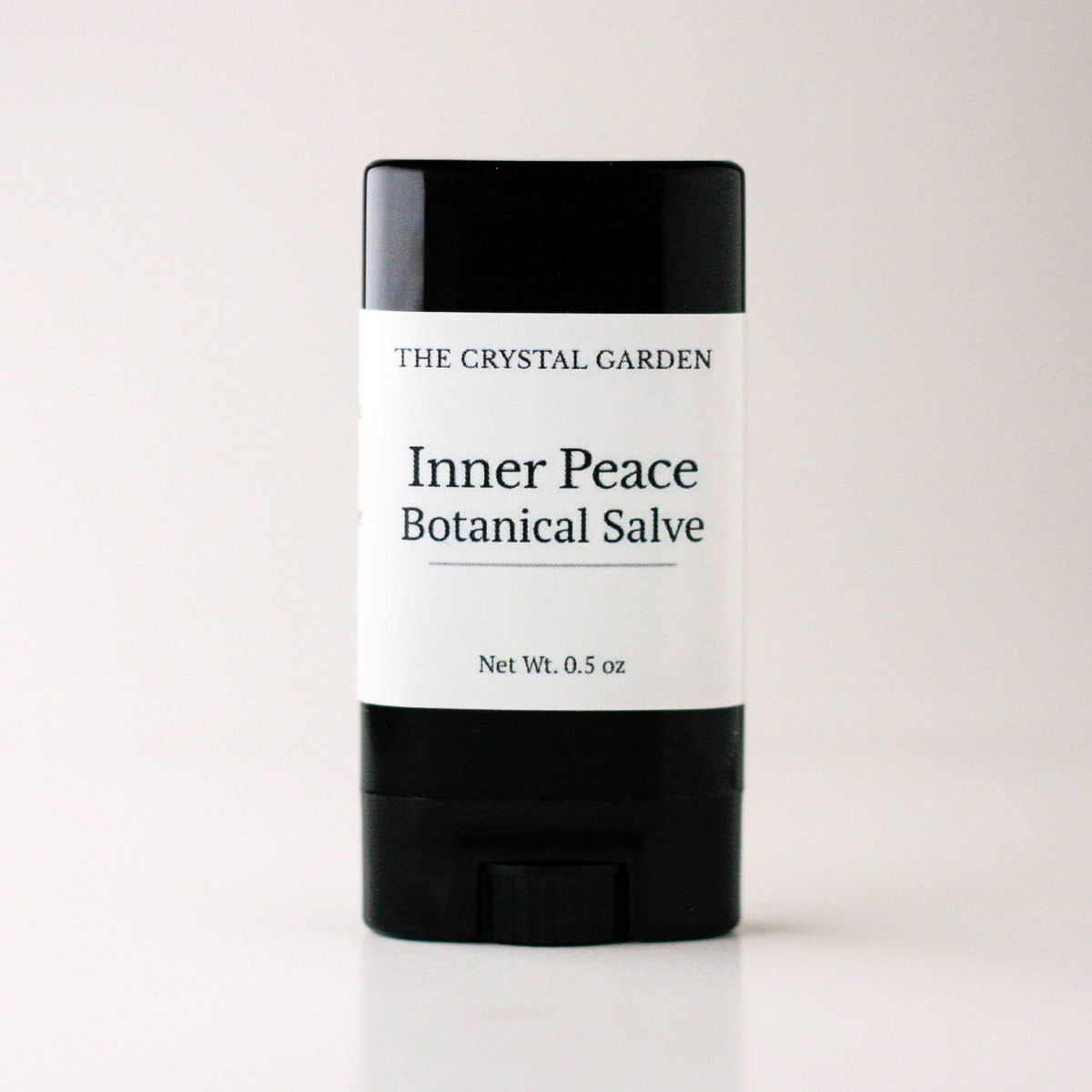 A single stick of salve from The Crystal Garden, specifically Inner Peace Botanical Salve, in a black plastic container with a white paper label, set against a plain light background.