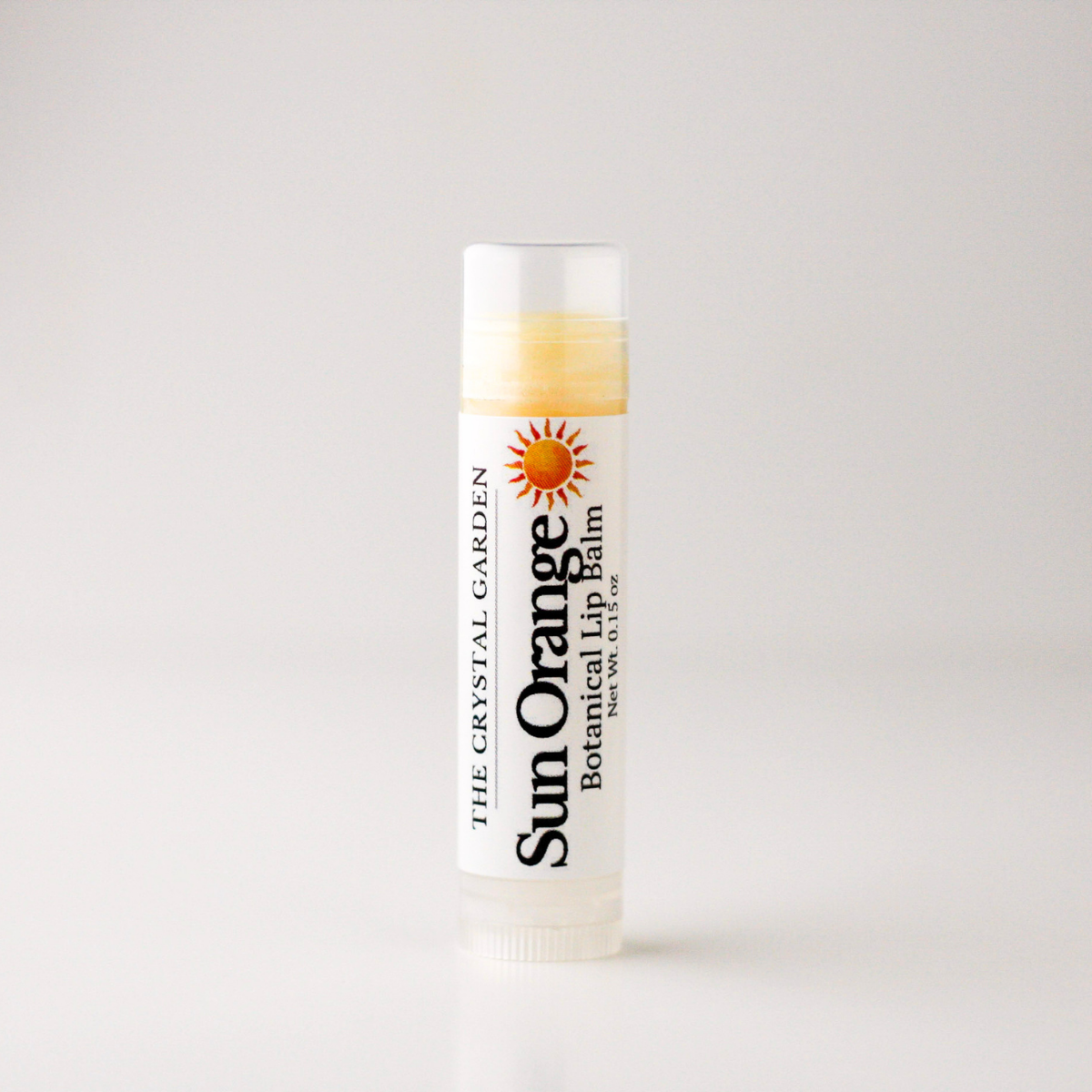 A single tube of Sun Orange Botanical Lip Balm by The Crystal Garden, standing upright on a clean white surface against a light gray background.