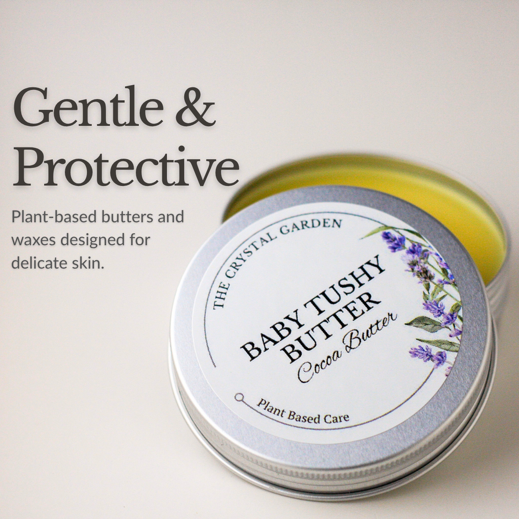 An open round silver tin of Baby Tushy Butter Cocoa Butter by The Crystal Garden, showing the pale yellow balm inside, with a white label featuring purple lavender illustrations. The image includes marketing text on the left reading 'Gentle & Protective'.