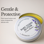An open round silver tin of Baby Tushy Butter Cocoa Butter by The Crystal Garden, showing the pale yellow balm inside, with a white label featuring purple lavender illustrations. The image includes marketing text on the left reading 'Gentle & Protective'.