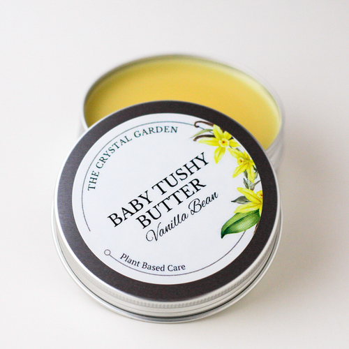 An open silver metal tin of Baby Tushy Butter in Vanilla Bean, showing the pale yellow balm inside, with the lid placed in front displaying a white label with black text and yellow flower illustrations. The brand 'The Crystal Garden' is visible on the label.