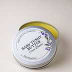 A round silver metal tin of Baby Tushy Butter Cocoa Butter by The Crystal Garden, with the lid off to reveal the yellow balm inside, set against a plain light beige background.