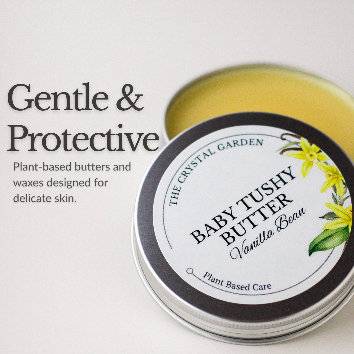 A round metal tin of Baby Tushy Butter in Vanilla Bean, shown open with pale yellow balm visible inside. The lid has a white label with black text and yellow flower illustrations.